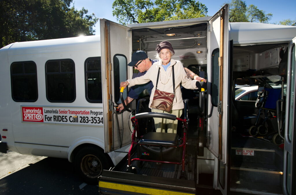 An elderly woman with a walker is being assisted by a man while exiting a senior transportation program minibus.