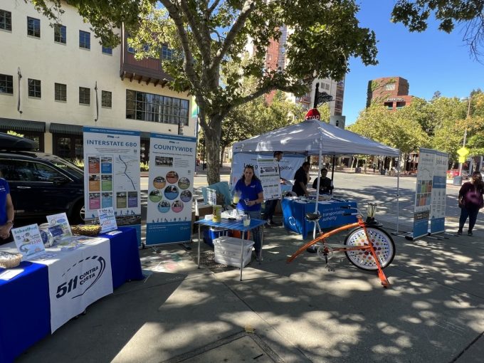 Outdoor event with booths promoting transportation initiatives and programs by the Contra Costa Transportation Authority, including Interstate 680 improvements and countywide services, with staff members and informational displays.