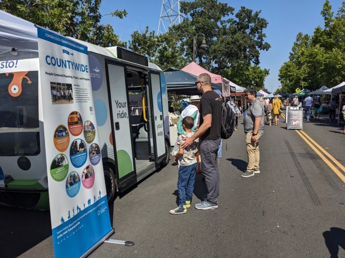 People examining an autonomous shuttle and information display about people-centered planning projects at an outdoor fair under sunny weather.