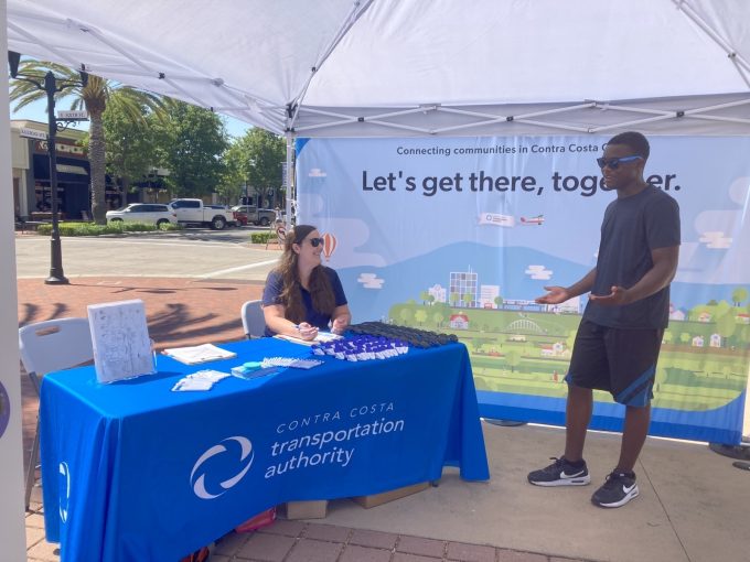 A man speaks with a woman sitting at a booth with a blue tablecloth from the Contra Costa Transportation Authority under a canopy, with a banner that says, "Let's get there, together."