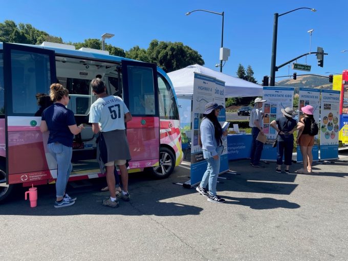 People at an outdoor exhibit featuring a pink autonomous vehicle and informational booths about highways and transportation.